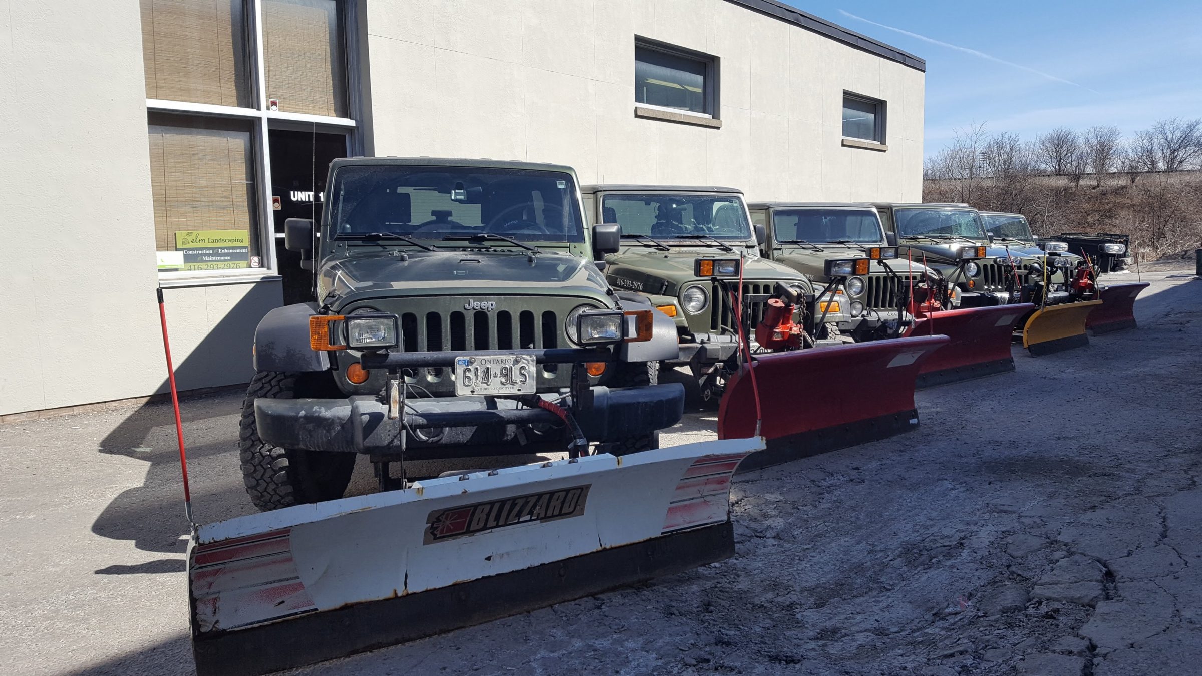 Four jeeps with snowplows parked outside building.