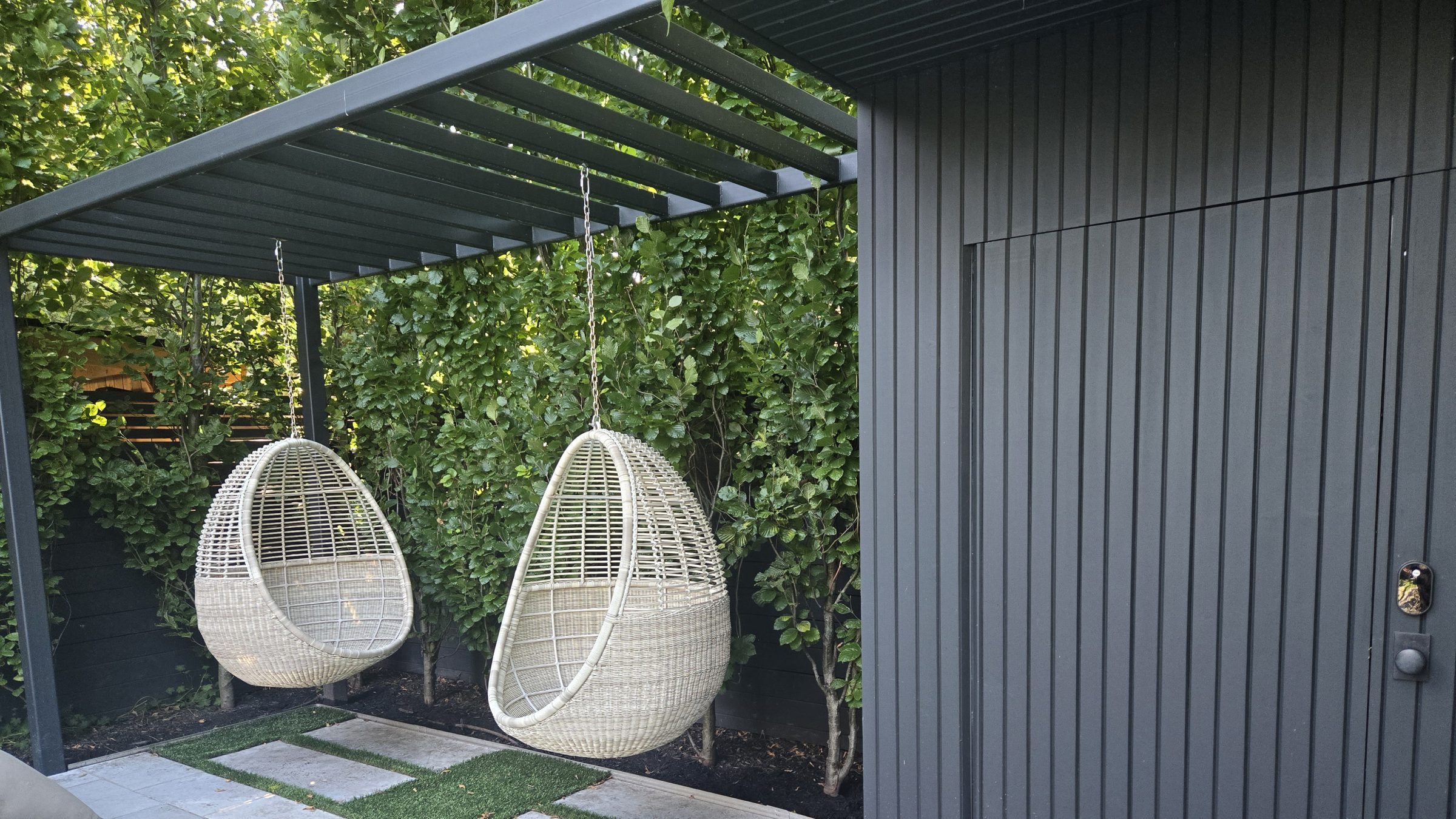 Outdoor egg chairs under pergola with leafy backdrop.